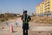 Man in uniform holding gun and standing on dirt road in front of building (© Thomas Peter/Reuters)